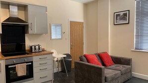 Un espace de vie compact comprenant un coin cuisine, un canapé gris avec des coussins rouges, une porte en bois et un tableau encadré au mur.