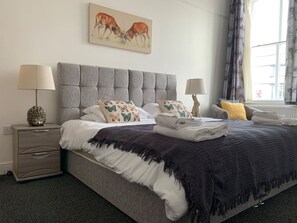 Una cama bien hecha con un cabecero gris, una mesita de noche con lámpara, y una ventana con cortinas.