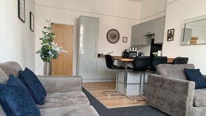 Ett modernt vardagsrum med en grå soffgrupp, ett soffbord och en matplats, med ett kök synligt i bakgrunden.