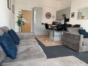 Un salon moderne avec un canapé gris, une table basse et un coin repas, la cuisine étant visible au fond.