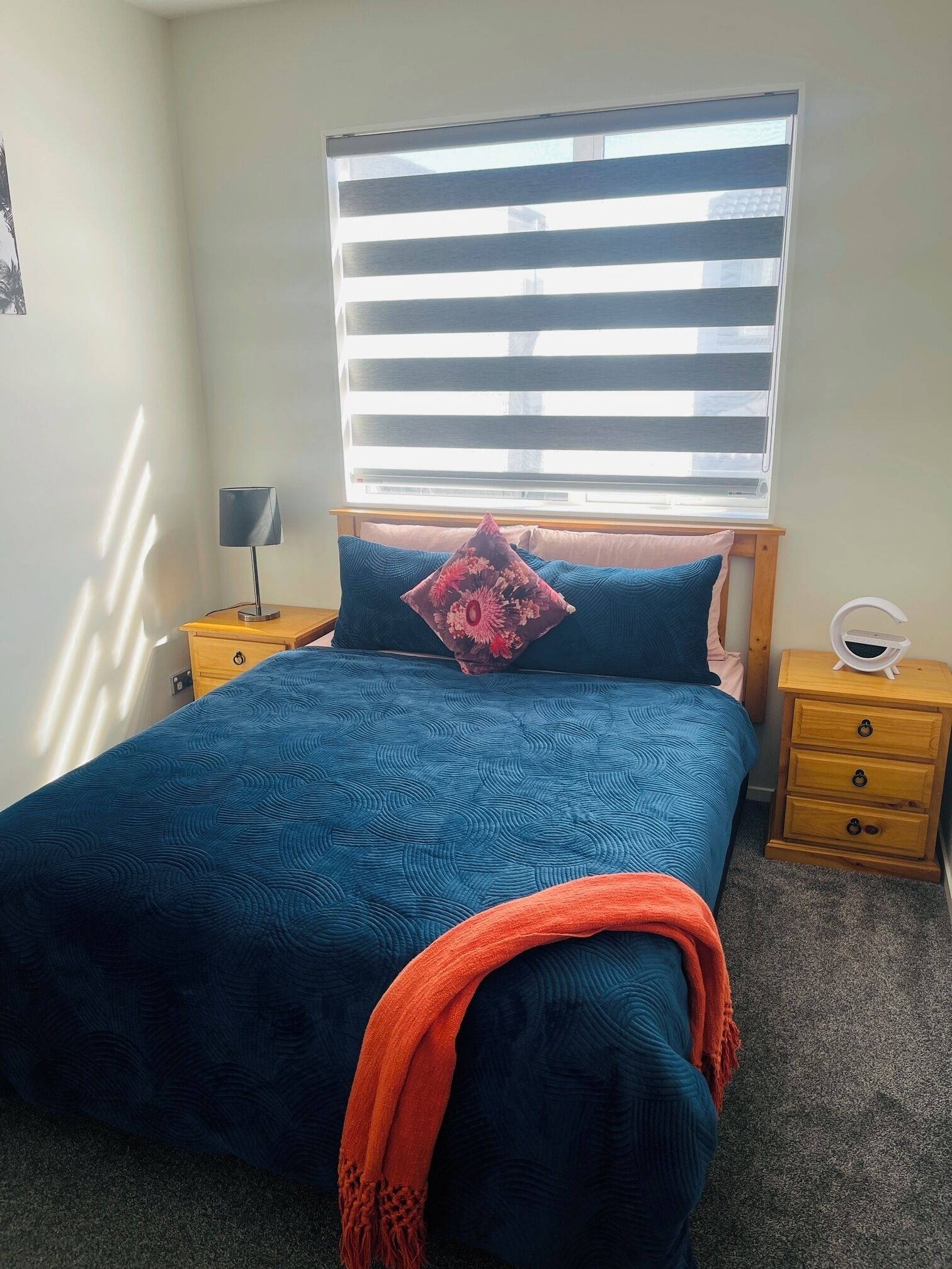 Une chambre à coucher comprenant un lit, une table de chevet, une lampe, une fenêtre avec des stores et un luminaire fixé au mur.