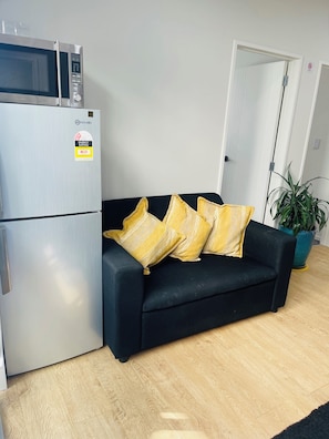 Un espacio habitable compacto con kitchenette, que incluye un microondas y un refrigerador, un sofá de color oscuro con almohadas amarillas, una planta en maceta y piso de madera.