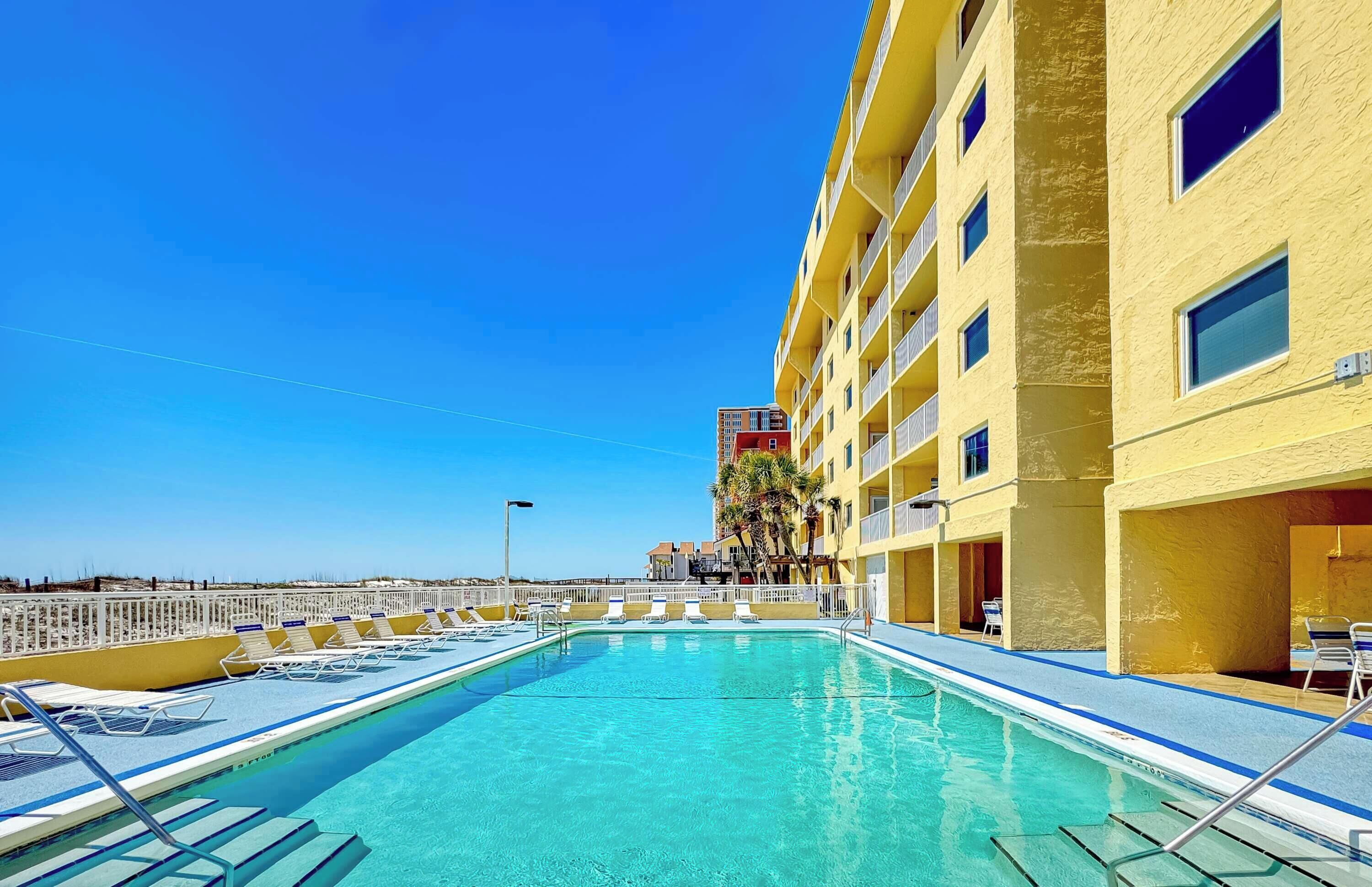 Una piscina con sillas de descanso, un edificio amarillo y un cielo azul despejado.