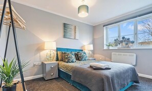 Un dormitorio con cama, lámparas de noche, radiador, ventana con vistas y una lámpara colgante.