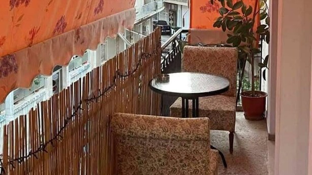 Un balcón con cortinas florales anaranjadas, una mesa redonda pequeña, dos sillas con estampado y una planta en maceta, con vista a un edificio al fondo.