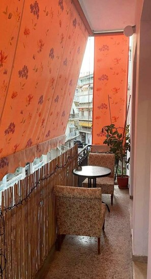 A balcony with orange floral curtains, a small round table, two patterned chairs, a potted plant, and a view of a building in the background. - Apartment Sleeps 4 Near Acropolis Museum (Athens)
