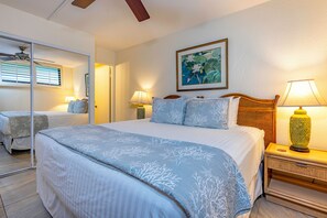 A bedroom with a large bed, two bedside tables with lamps, a ceiling fan, a framed picture on the wall, and a mirror reflecting the room. - Kihei Surfside #311 - Charming Oceanfront Condo with AC: Your Perfect Retreat (Kihei, Maui)