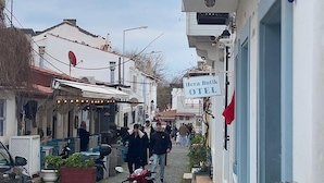 A narrow street with shops and outdoor seating, cobblestone pavement, and a sign for 'Hera Butik' on the right. - HERA BUTİK OTEL (Bozcaada)