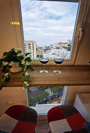 A balcony with two wine glasses, a potted plant, and a view of a cityscape. - Exclusive Designer Apartment | Stunning Sea Views & Walk to Beach (Benalmádena)
