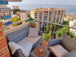 A balcony with wicker furniture, a view of the city, and a sign that says 'LIFE is Better at the BEACH.' - Exclusive Designer Apartment | Stunning Sea Views & Walk to Beach (Benalmádena)