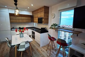 A modern kitchen with wooden cabinets, a dining table with patterned chairs, a red and black chair, a small bar area with stools, a clock, and a window with a city view. - Exclusive Designer Apartment | Stunning Sea Views & Walk to Beach (Benalmádena)
