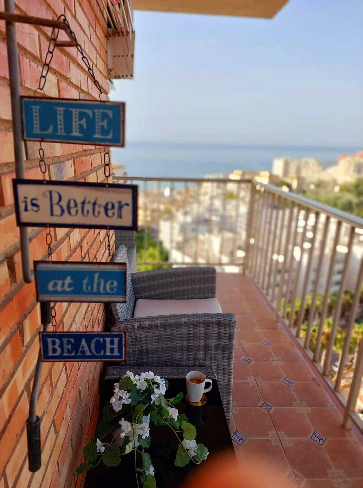 Een balkon met een rieten bank, een koffiekop en een bordje met de tekst 'Het leven is beter aan het strand'.