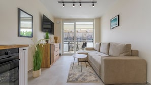 A modern living room with a sofa, a small table, a kitchen area with a stove, and a balcony with a view. - Apartamenty Sun&Snow Baltic Sun II (Sztutowo)