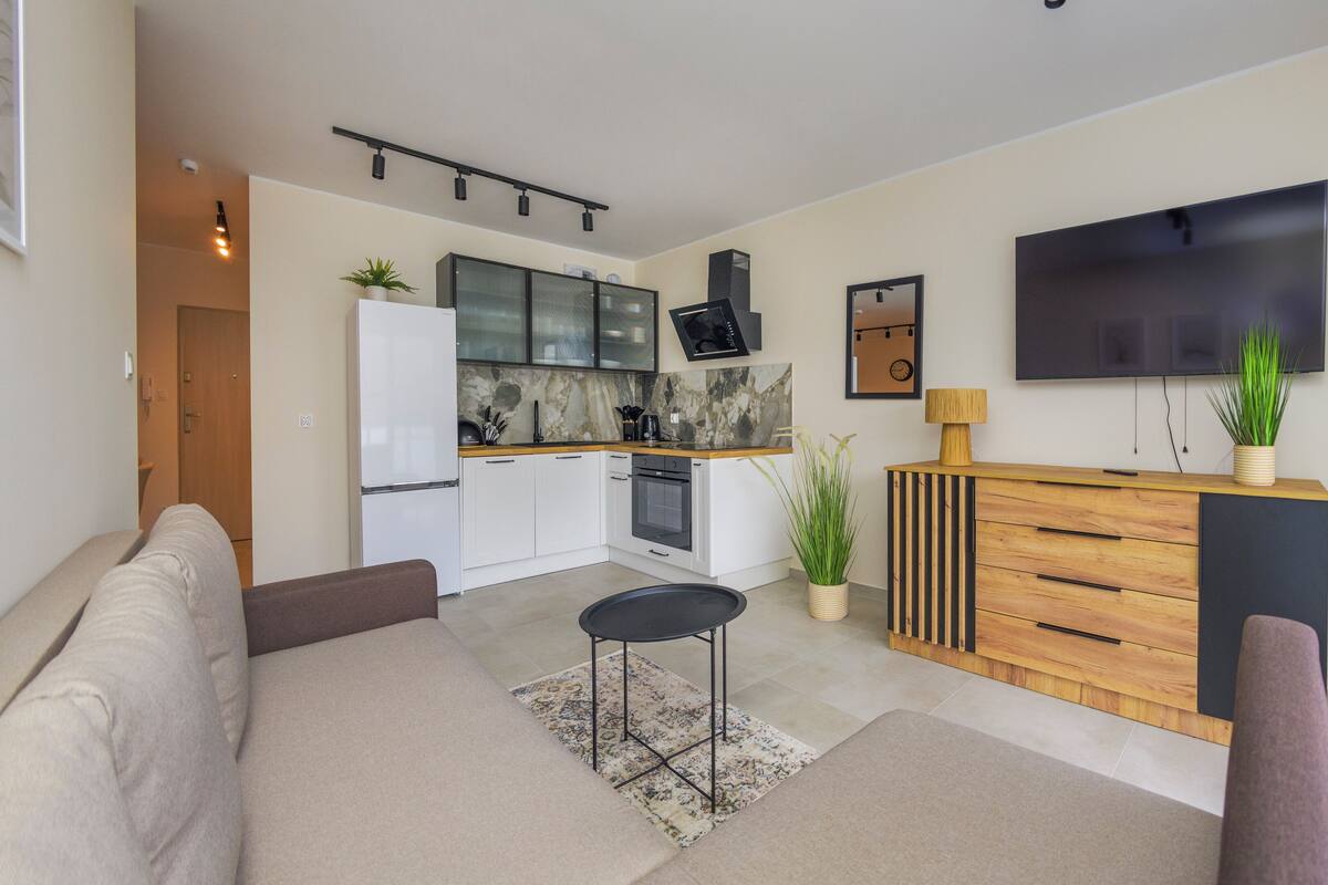 A modern kitchen with a built-in oven, a dining area with a sofa and a small table, and a flat-screen TV mounted on the wall.