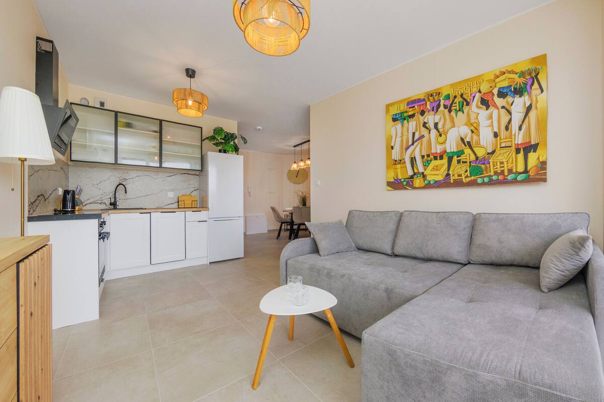 A modern living room with a grey sofa, a small white table, and a kitchen in the background.