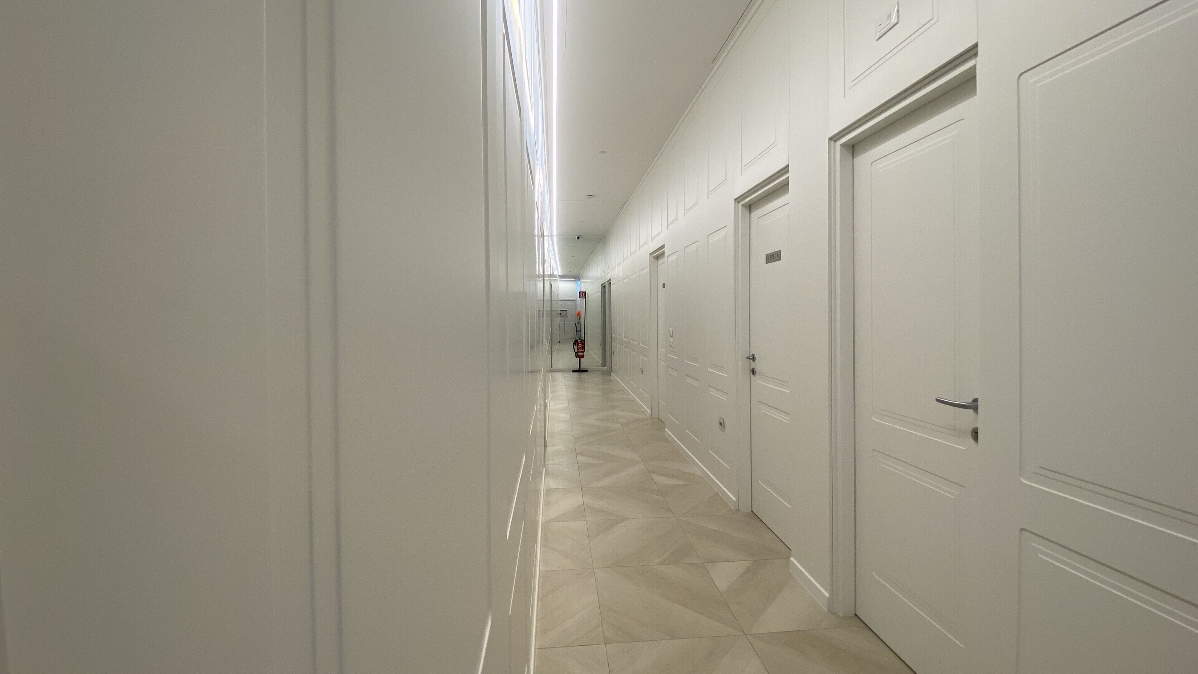 A long, narrow hallway with white doors on one side and a fire extinguisher mounted on the wall.