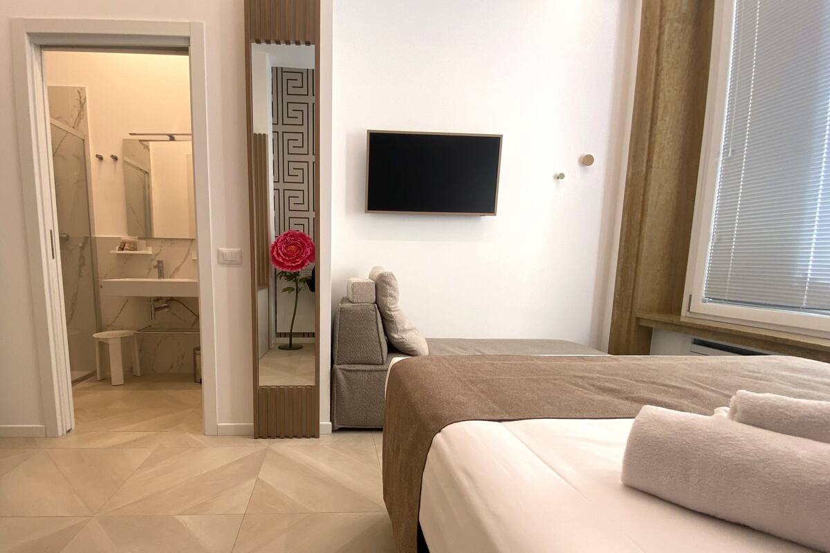 A hotel room with a bed, a TV, a bathroom with a mirror, and a window.