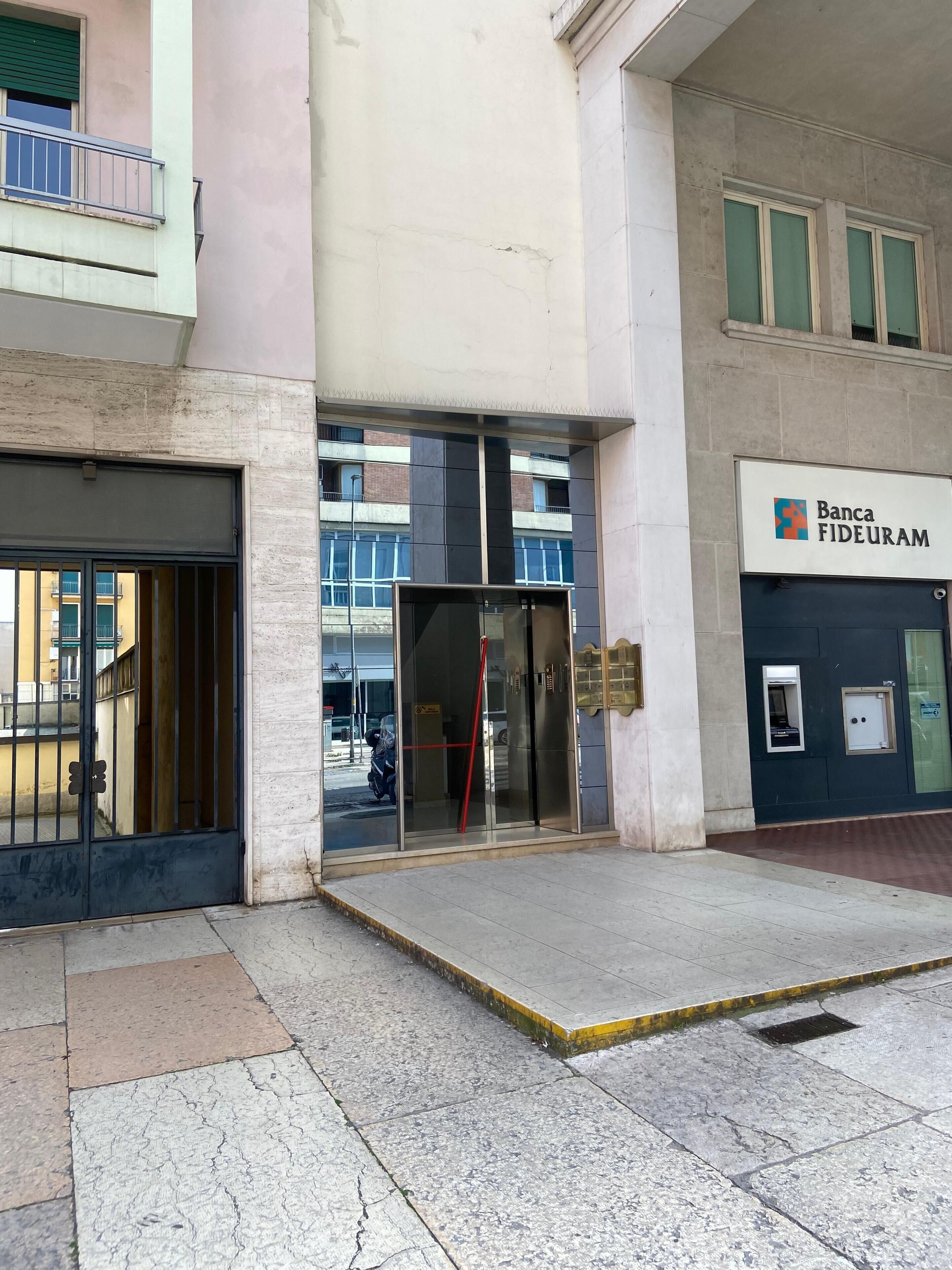 Une entrée d’immeuble avec une porte en verre, un portail métallique et une enseigne indiquant « Banca Fideuram ».