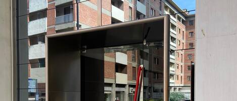 A modern elevator with a glass enclosure, a red pole inside, and a view of a multi-story building with balconies in the background.