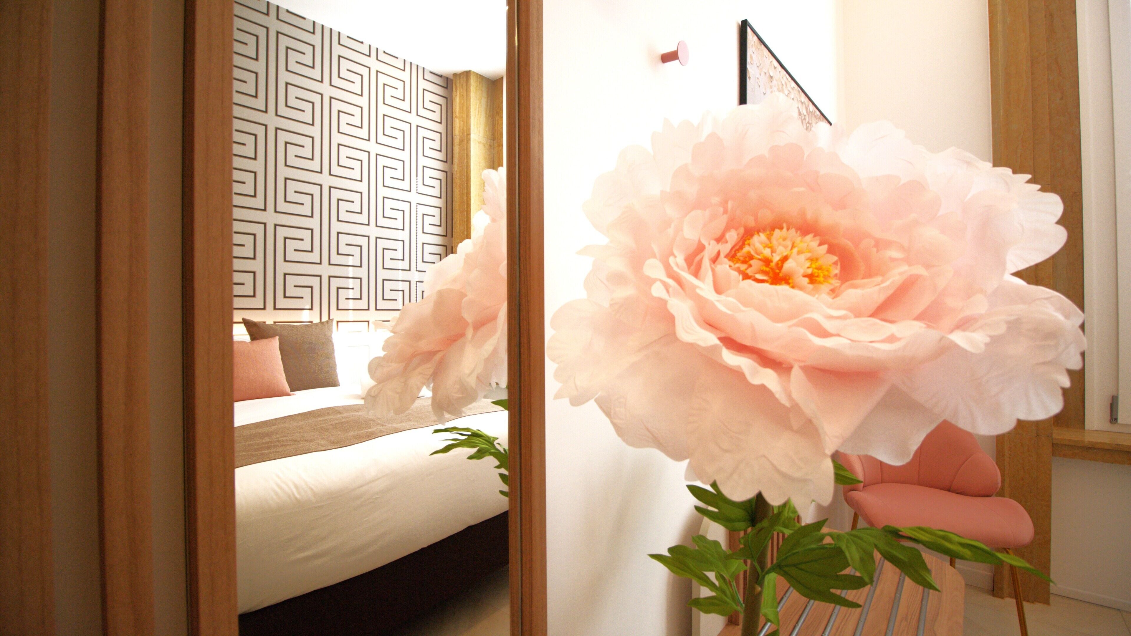 A bedroom with a bed, a mirror, a pink flower, and a geometric wall pattern.