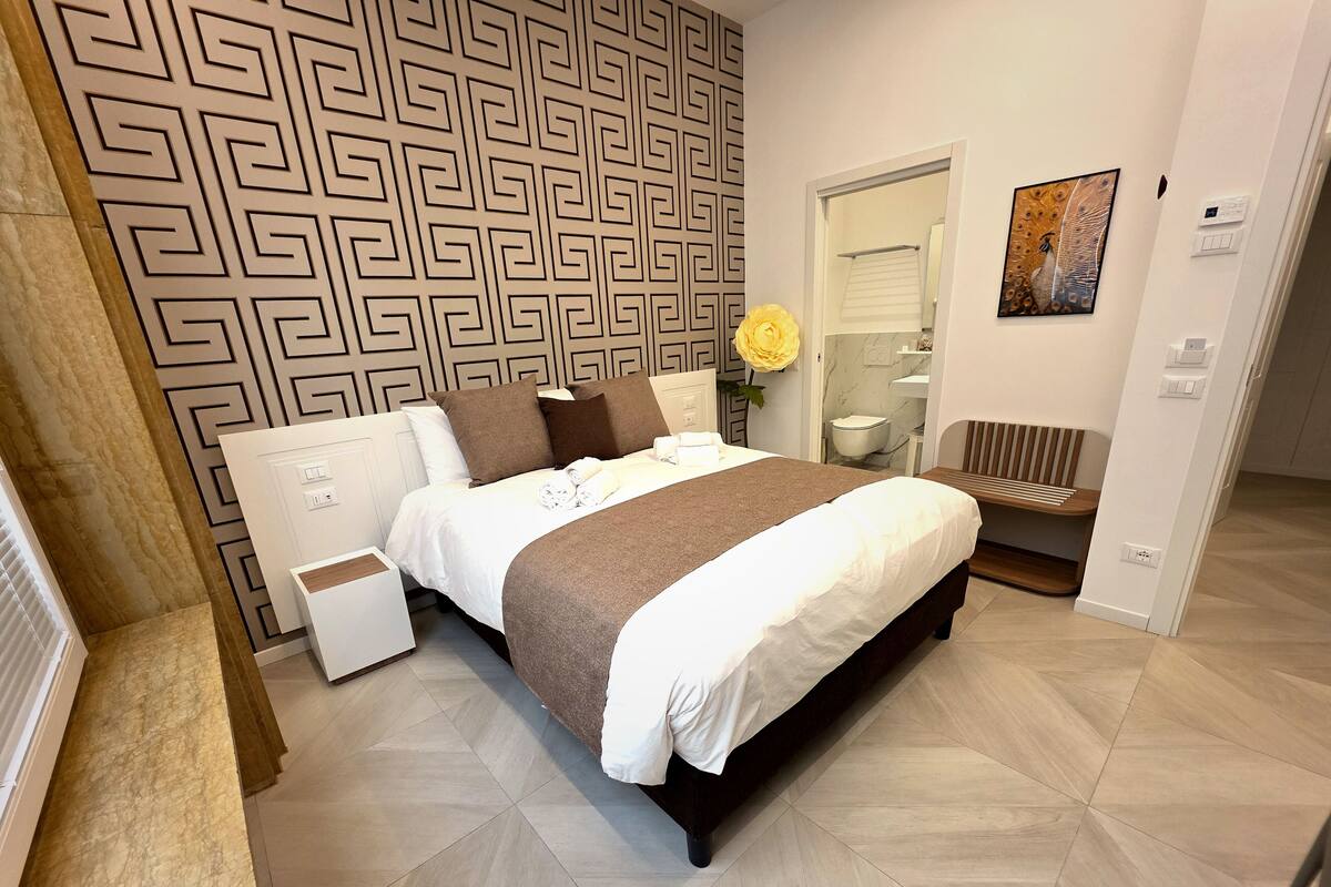 A modern hotel room with a large bed, a bedside table, a mirror, a bathroom with a toilet and sink, and a decorative wall pattern.