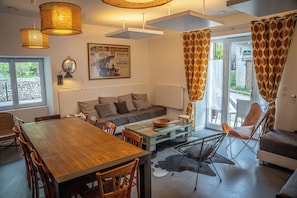Uma sala de estar moderna com mesa de jantar de madeira, sofá, cadeiras, mesa de centro de vidro e uma janela grande com cortinas.