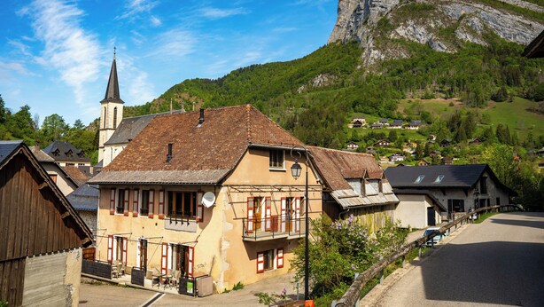Ein traditionelles europäisches Dorf mit einer Kirche und einem markanten Berg im Hintergrund, verbunden durch eine gepflasterte Straße.