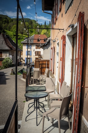 A balcony with wicker chairs and a table, overlooking a street with buildings and a mountain in the background.