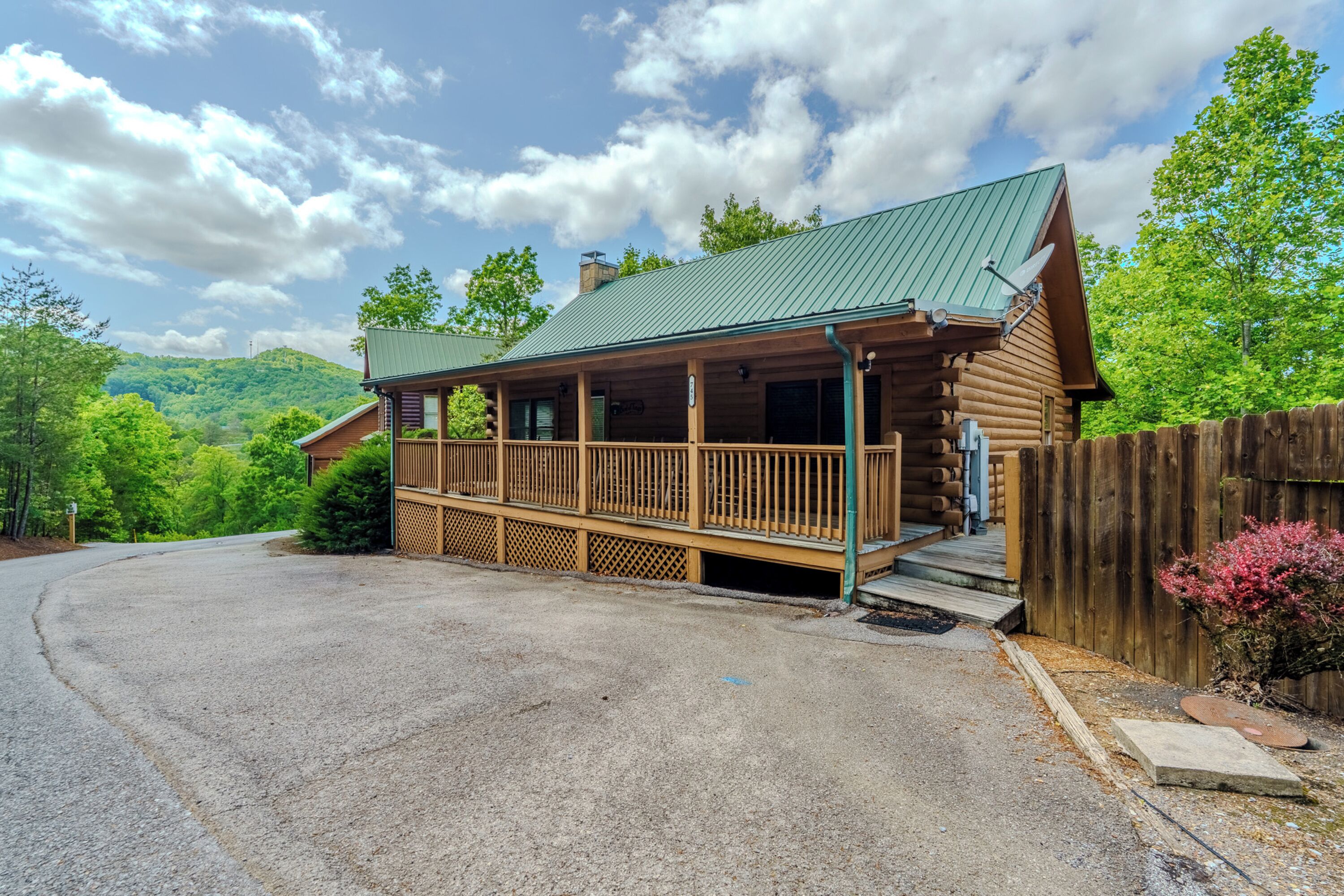 A log cabin with a porch, a green roof, and a view of a forested hill.
