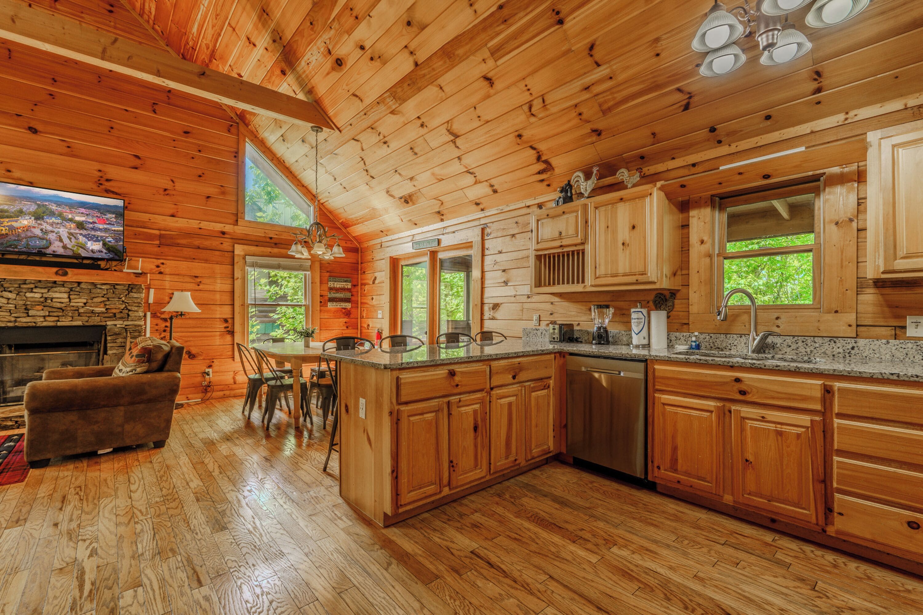 A wooden cabin interior with a stone fireplace, wooden dining area, kitchen with granite countertops, and large windows.