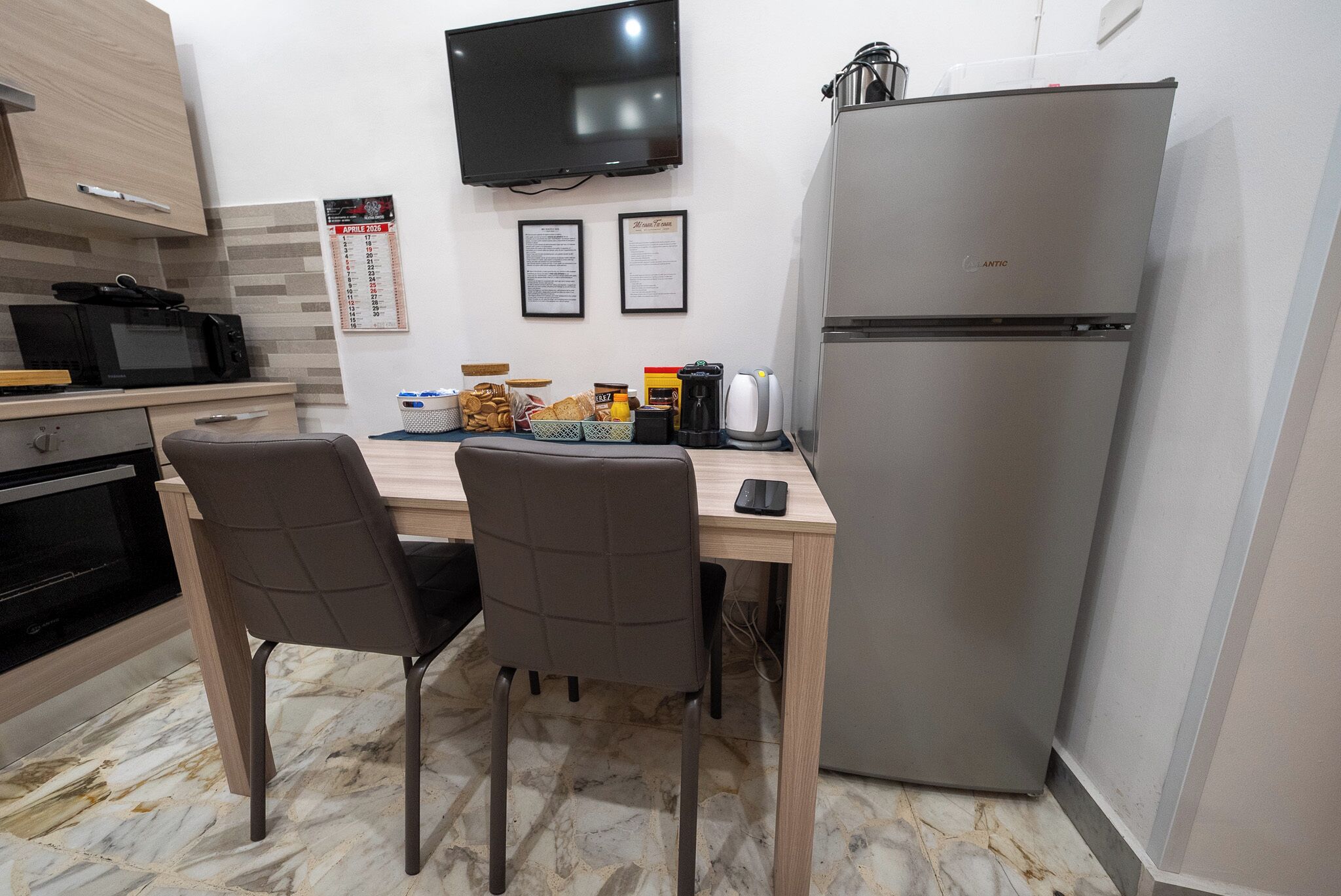 A kitchen with a wooden table, two chairs, a refrigerator, a microwave, and a TV mounted on the wall.