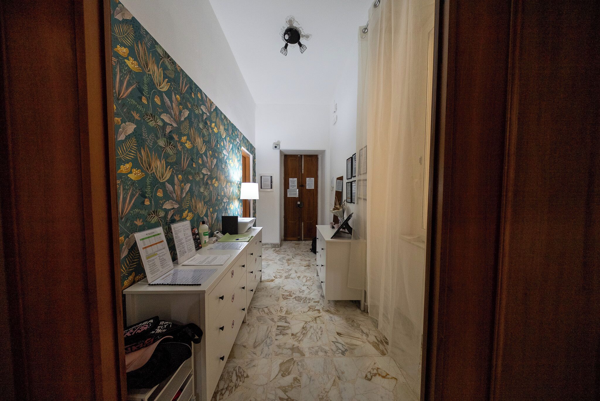 A hallway with a patterned wall, a desk with papers, a lamp, and a door at the end.