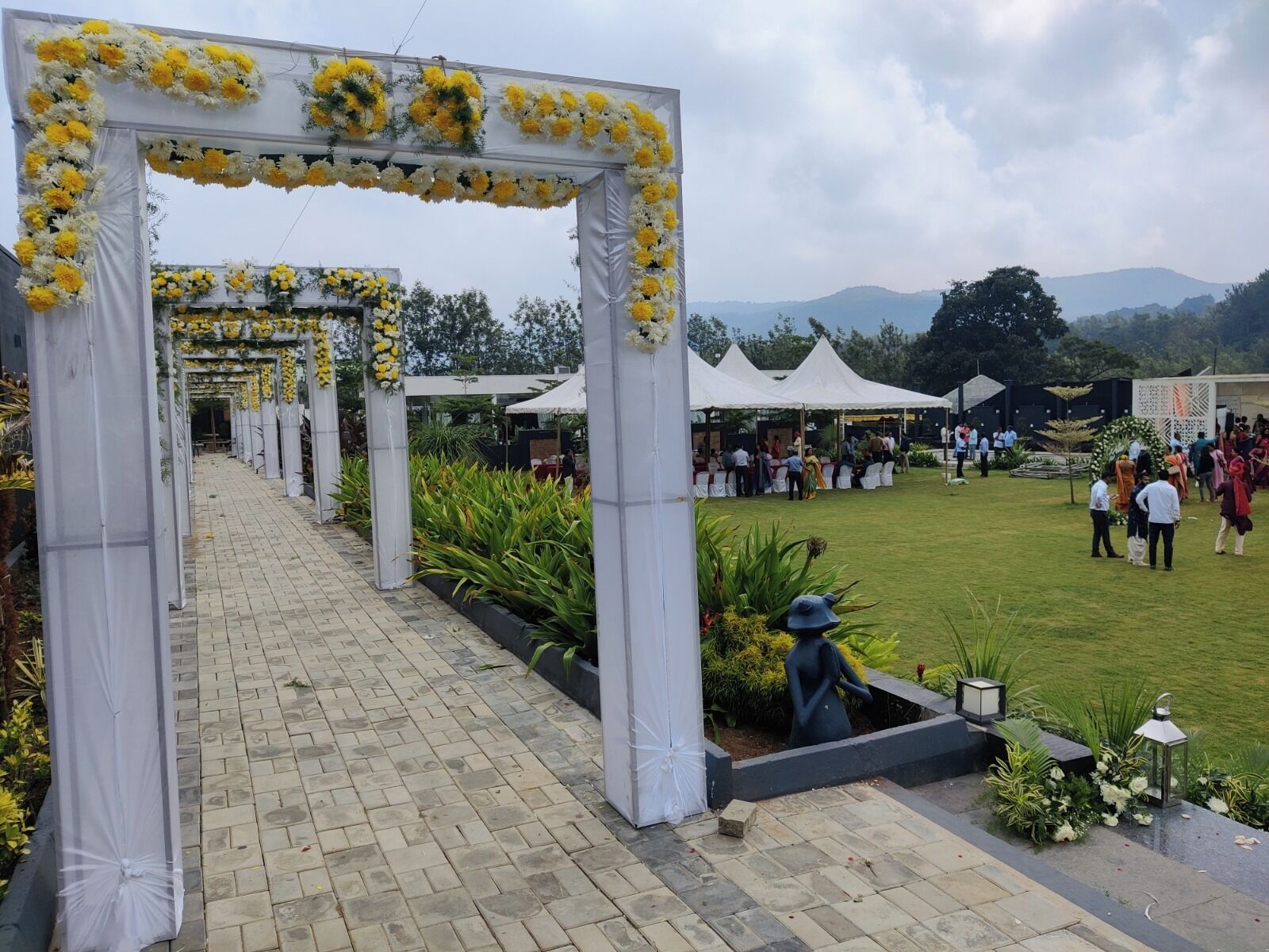 A pathway lined with white arches adorned with yellow and white flowers, leading to a grassy area with tents and people.