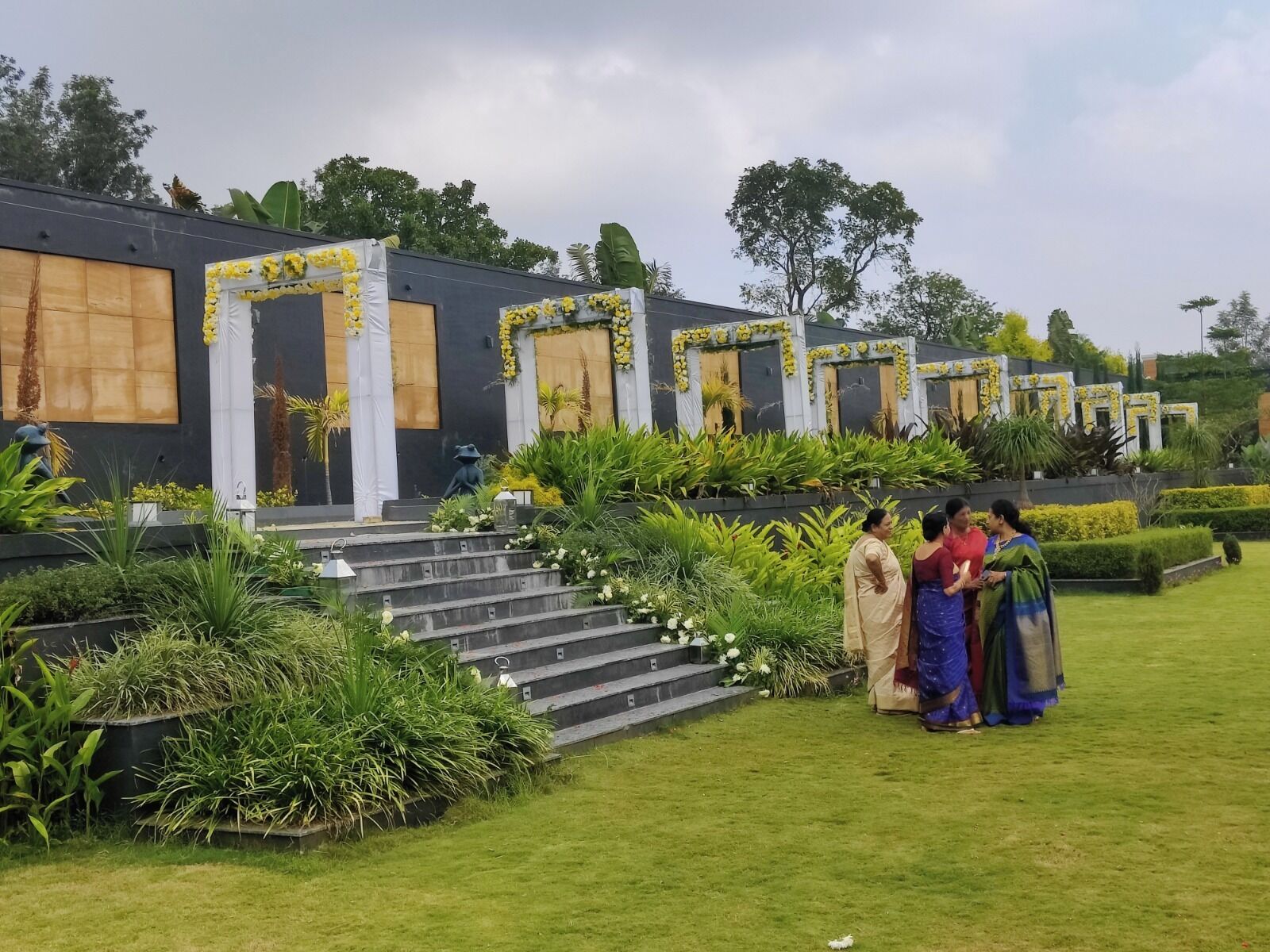 A building with multiple doors decorated with yellow flowers, surrounded by well-maintained greenery and a small staircase leading to the entrance.