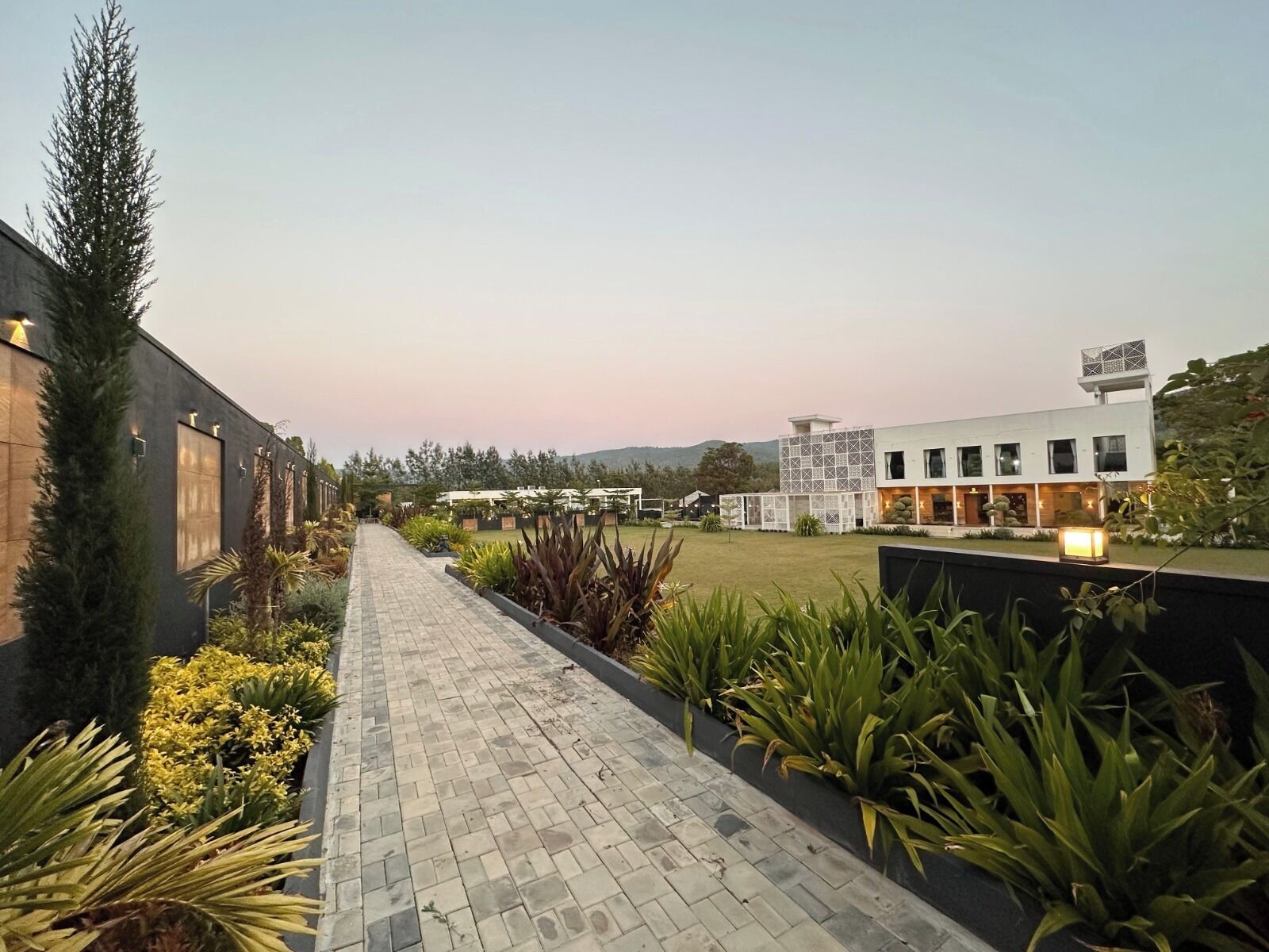 A pathway leading to a modern building with a flat roof and a small tower, surrounded by landscaped gardens and a clear sky.