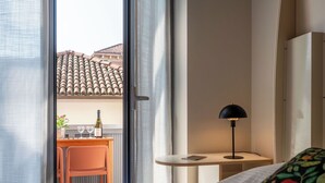 Hotellihuone, jossa on sänky, työpöytä tuolilla, lamppu ja näkymä rakennukseen, jonka katto on päällystetty tiilillä.