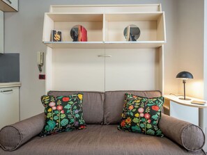 A sofa with floral cushions, a white shelf with books and decorative items, a small table with a lamp, and a wall-mounted phone. - Cosy apartment for 3 people with WIFI, A/C, TV and balcony (Alba)