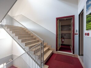 Eine Treppe mit Metallgeländer, einer Teppichlandung in Rot und einer Tür zu einem Raum mit Feuerlöscher und einer wandmontierten Steuerung.