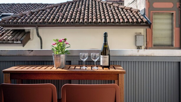 Een houten tafel met een fles wijn, twee glazen en een bloempot op een balkon met een betegeld dak en een gebouw op de achtergrond.