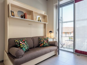 Ein Wohnzimmer mit einer Couch, einem kleinen Tisch mit Lampe, einer Schiebetür und einem Balkon mit Tisch und Stühlen.