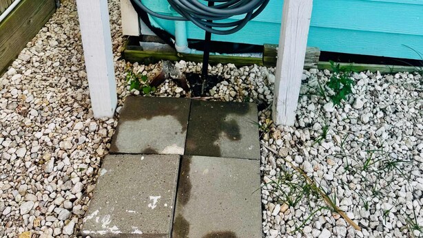 Un tuyau d’arrosage enroulé contre un mur en bois turquoise, une sonnette métallique et un chemin en béton.