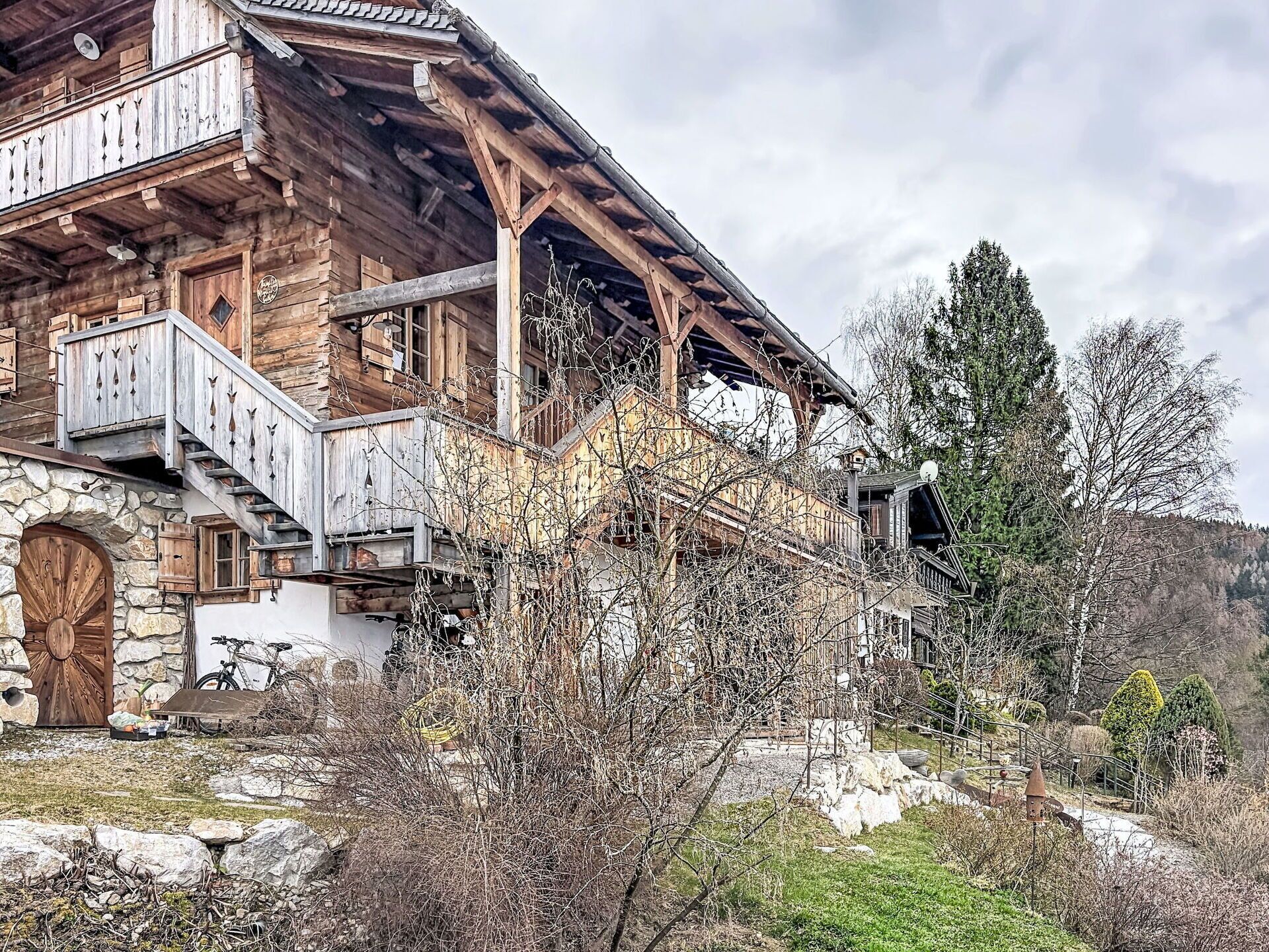 A wooden chalet with a stone entrance, wooden balconies, and a staircase leading to the upper floor.