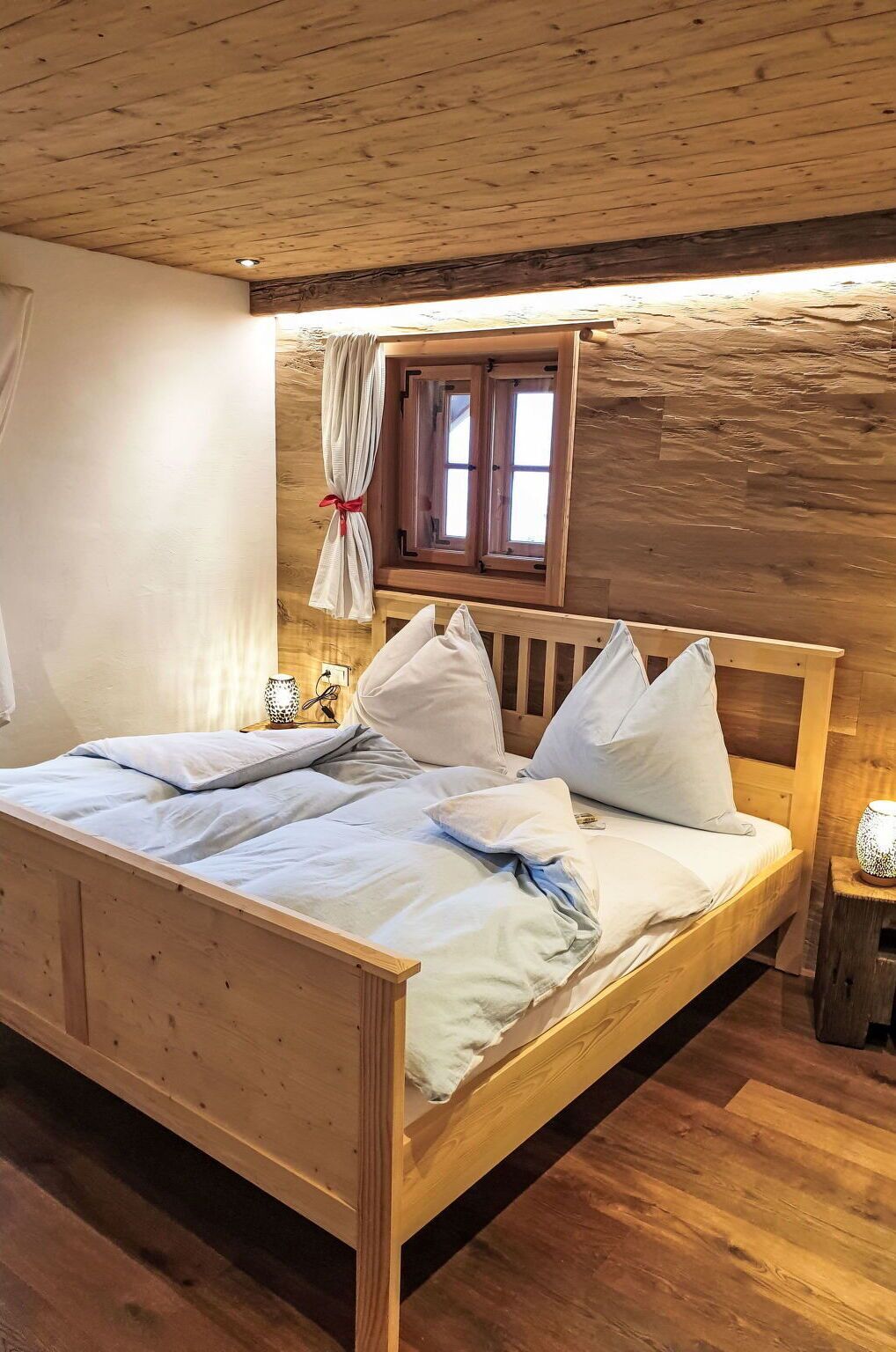 A wooden bed with white bedding, a window with curtains, and a small table with a lamp.