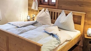Ein hölzernes Bett mit weißer Bettwäsche, ein Fenster mit Vorhängen und ein kleiner Tisch mit einer Lampe.