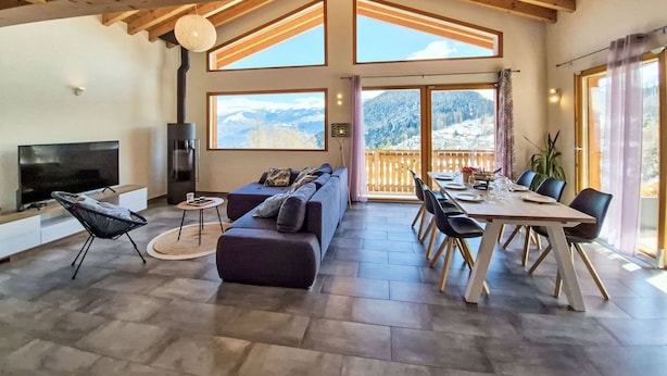 Ein geräumiger Wohnbereich mit Kamin, einer Couch, einem Esstisch mit Stühlen und großen Fenstern mit Blick auf die Berge.