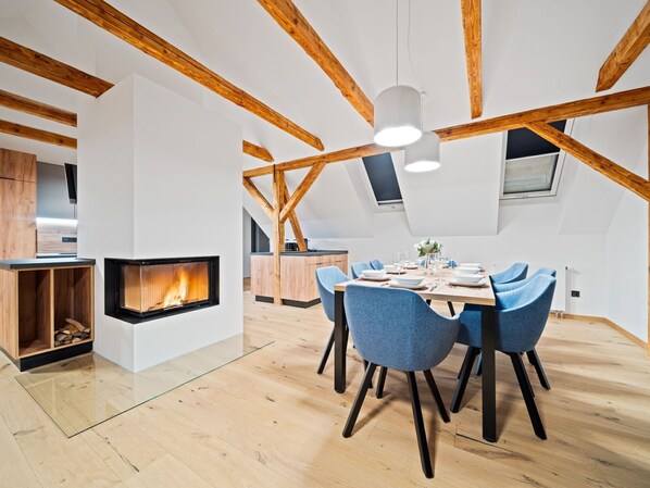 A modern loft with exposed wooden beams, a fireplace, a dining area with blue chairs, and a kitchen with wooden cabinets.