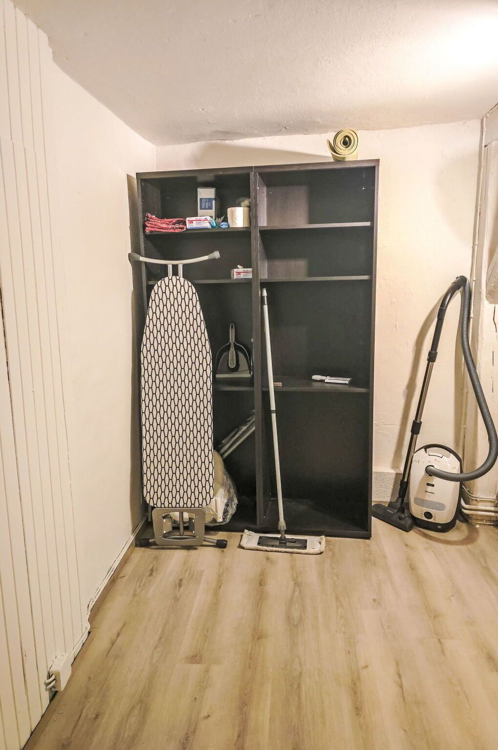 Una habitación con piso de madera, un estante negro con una plancha blanca, un aspirador y un trapeador.