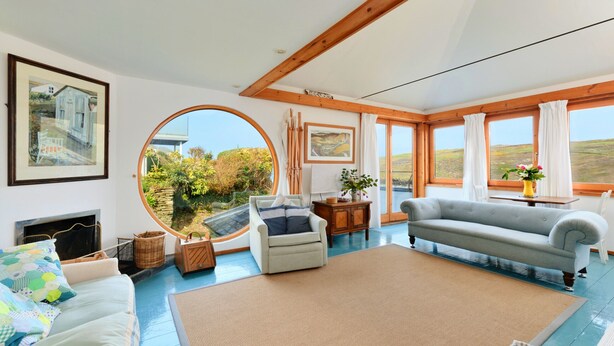 A spacious living room with a large round window, a sofa, a fireplace, and a painting on the wall. - The Cuddy (Porthcothan Bay)