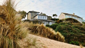 A coastal house with a sandy foreground, dense vegetation, and a clear sky. - The Cuddy (Porthcothan Bay)