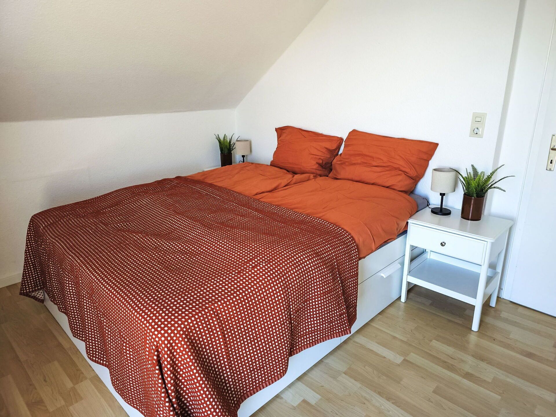 A bedroom with a bed, a nightstand, a lamp, and a potted plant.